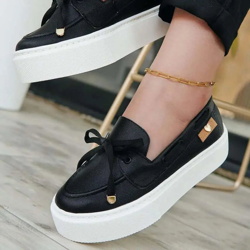Mocasín negro de mujer con plataforma blanca, detalle de lazo y pequeño accesorio dorado en forma de corazón, calzado cómodo, versátil y moderno ideal para uso diario, fabricado con materiales de alta calidad que garantizan durabilidad y estilo.