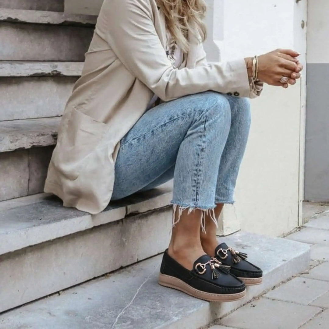 Mocasines negros para mujer con detalle de borlas y hebillas doradas, diseño cómodo y casual, suela antideslizante y ligera, ideales para uso diario y combinan con jeans y chaqueta beige, calzado elegante y versátil para estilo urbano contemporáneo.