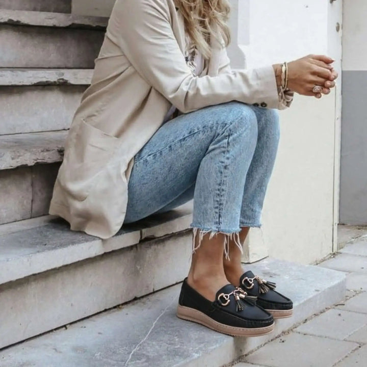 Mocasines negros para mujer con detalle de borlas y hebillas doradas, diseño cómodo y casual, suela antideslizante y ligera, ideales para uso diario y combinan con jeans y chaqueta beige, calzado elegante y versátil para estilo urbano contemporáneo.