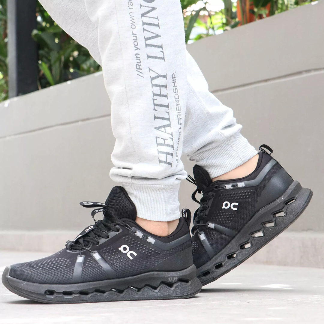 Vista lateral de tenis On negros para hombre con suela amortiguada CloudTec del mismo color y logo blanco. Combinados con un pantalón de sudadera gris claro con textos estampados.