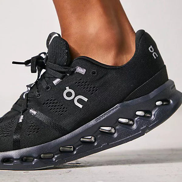Plano detalle de tenis On negros con suela de amortiguación CloudTec Phase del mismo color. Se observa el logo blanco de la marca sobre la malla transpirable y el calzado en posición de dar un paso