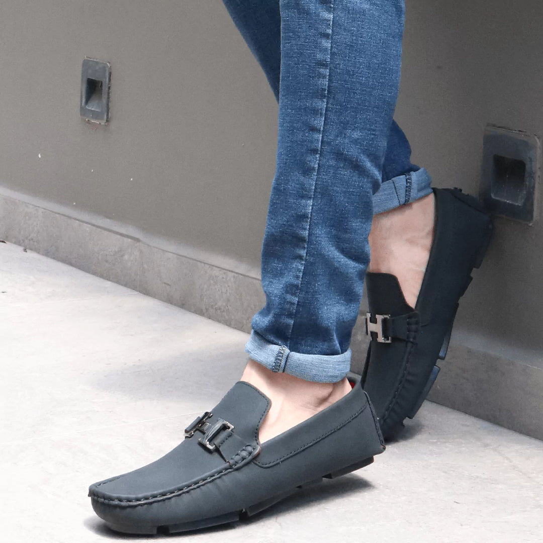 Hombre joven sentado usando zapatos Heritage negros tipo mocasín, con suela antideslizante, cómodos y versátiles para uso diario, combinados con pantalón jean azul y camiseta negra, en un ambiente moderno con plantas al fondo que resaltan un estilo casual y moderno ideal para la ciudad.