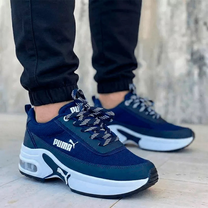 Tenis deportivos unisex de estilo robusto en color azul marino con plataforma blanca y cámara de aire visible. El calzado se apresenta en un modelo masculino con jogger negro, destacando un diseño moderno e imponente para el mercado colombiano