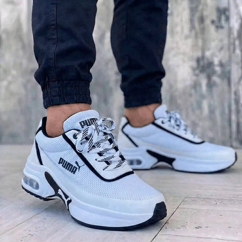 Tenis urbanos unisex blancos con detalles negros contorneados e suela de plataforma con amortiguación de aire visible. Fotografía enfocada en pies masculinos con pantalón jogger, ideal para un look versátil y sofisticado.