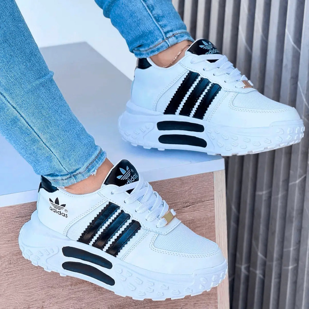 Zapatillas deportivas blancas con diseño moderno y tiras negras, suela gruesa y antideslizante tipo tractor para mayor comodidad y soporte, ideales para uso diario y actividades al aire libre, estilo casual y urbano perfecto para combinar con jeans ajustados.